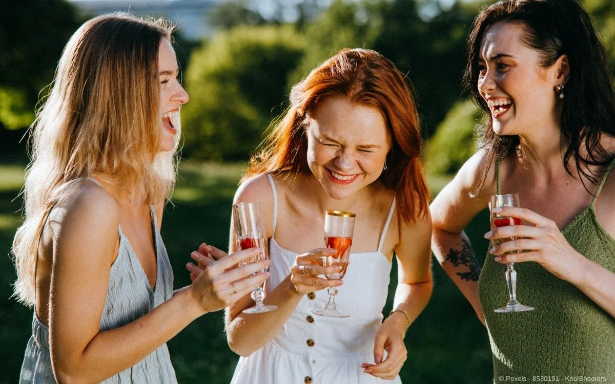 Drei fröhliche und ausgelassene Frauen die Prosecco auf einer schönen Wiese trinken